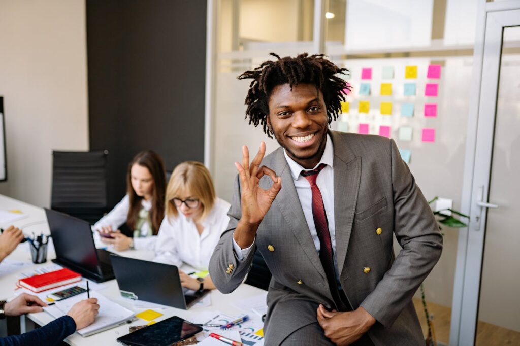 young man smiling at the camera doing an "ok" sign with team working in the background